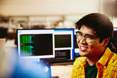 male smiling while sitting behind his desk, computer screens displaying programming code.