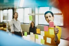 Two business women and a man in an office putting sticky notes on a window.